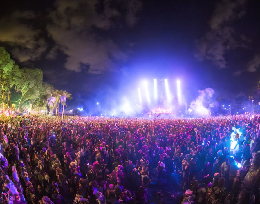 Nighttime outdoor music festival with large crowd, illuminated stage, trees, and purple sky.