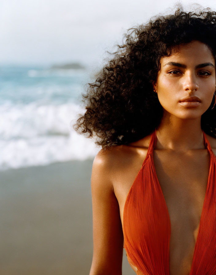 Curly-haired woman in orange halter top on beach with waves.
