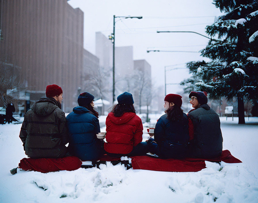 Six People in Winter Attire Chatting on Red Blanket in Snow