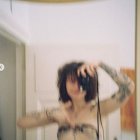 Tattooed person with short dark hair gazes into hazy mirror in softly lit room