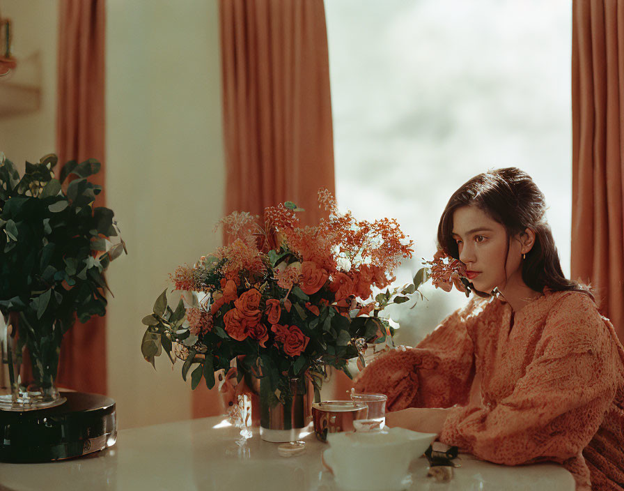 Person sitting at table with orange flowers, tea, and greenery in sunlit room