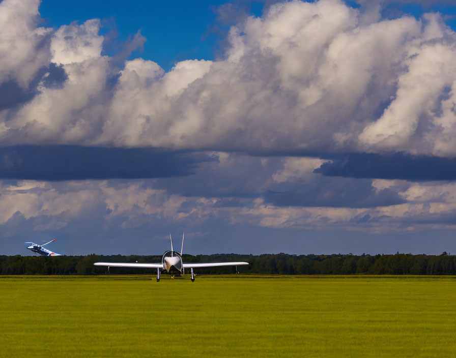 Glider soaring over lush field with dramatic cloudy sky and small aircraft taking off.