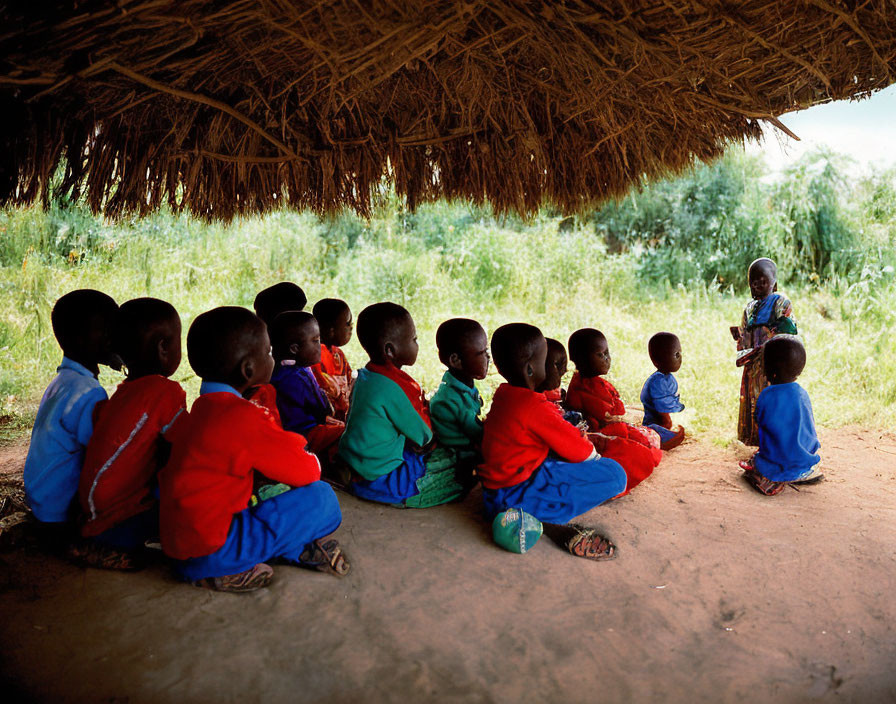 Young children in colorful attire listening to adult in thatched shelter