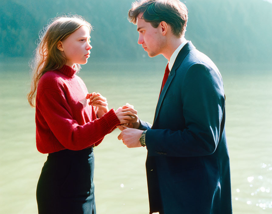 Two people holding hands by water and forest - one in red sweater, the other in dark suit