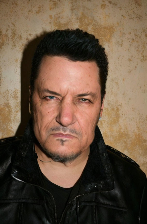 Stern man in black leather jacket with goatee and tattoo on beige backdrop
