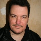 Stern man in black leather jacket with goatee and tattoo on beige backdrop