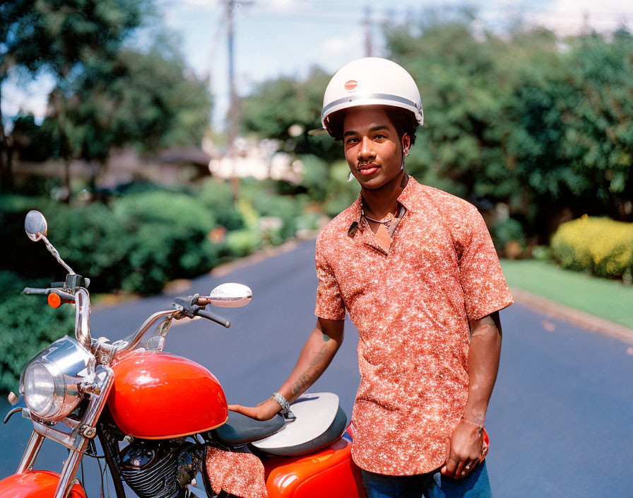 Young man with mustache in helmet and patterned shirt beside red motorcycle on suburban street