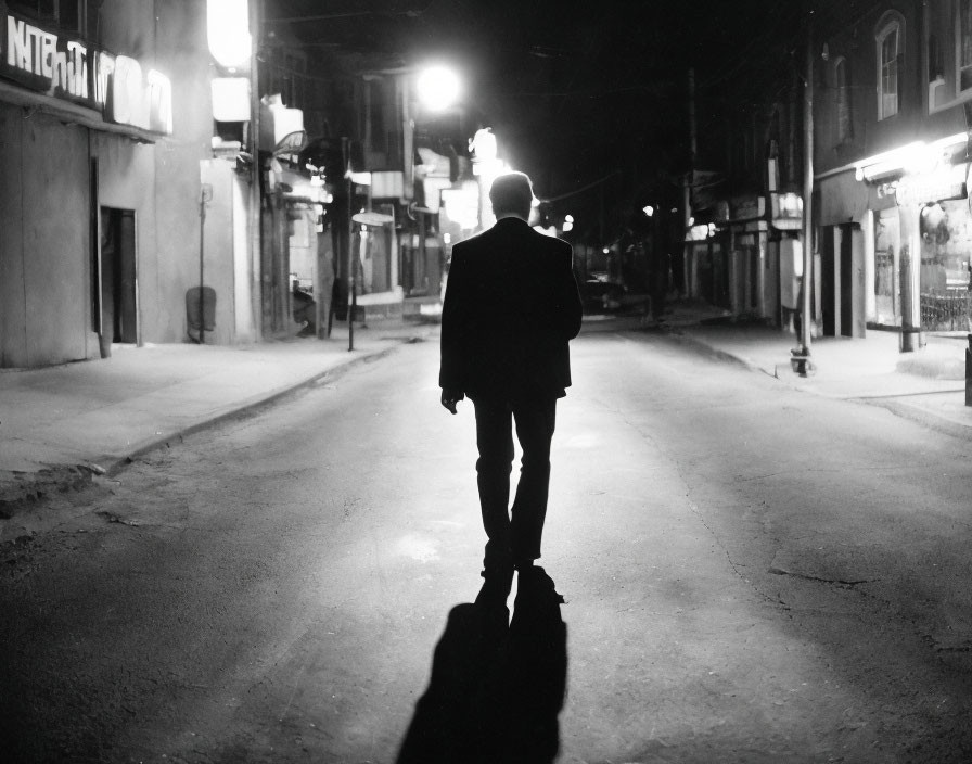 Person in suit walks down dimly lit street at night with glowing street lights and long shadows.