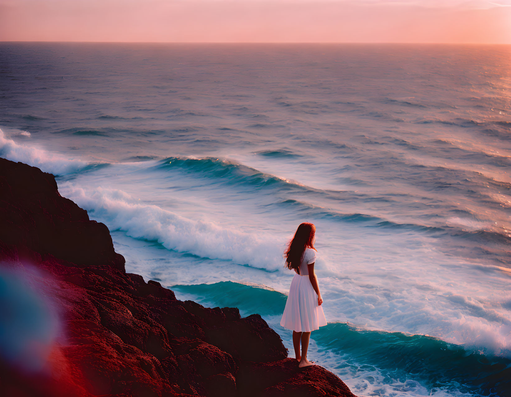 Woman in White Dress on Rocky Cliff Overlooking Ocean at Sunset