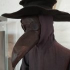 Cloaked figure in plague doctor mask at European building with clock tower.