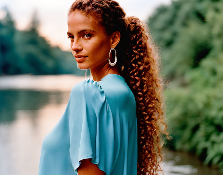 Curly-haired woman in blue blouse with hoop earrings by serene water.