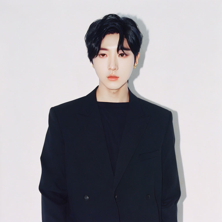 Black-haired individual in earring and dark blazer on white backdrop