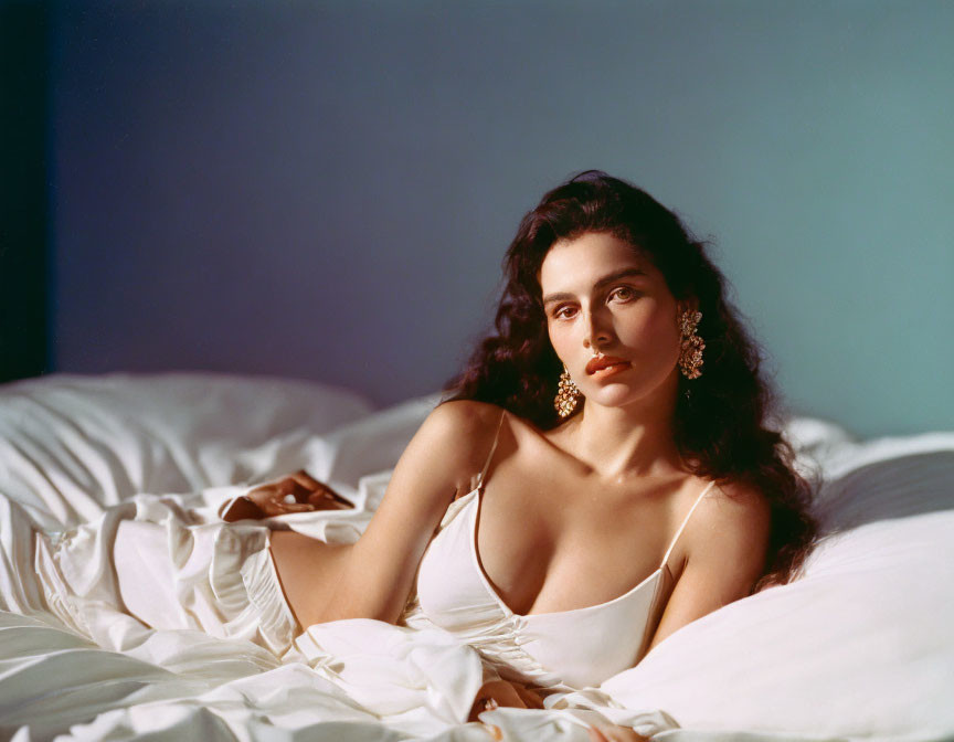 Woman in White Dress on Bed with Large Earrings in Dimly Lit Room