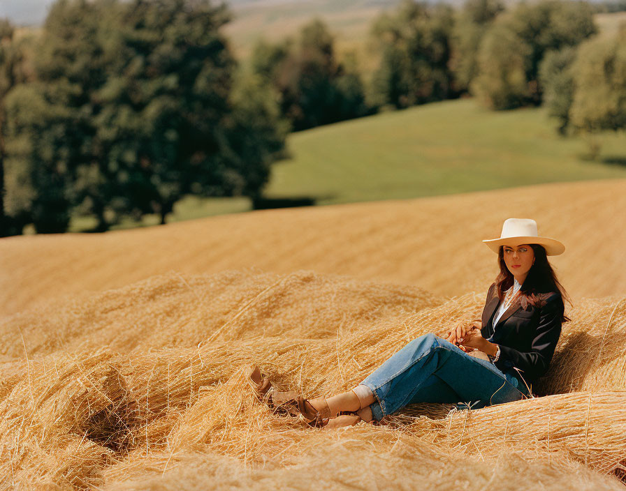 Woman in cowboy hat sitting in golden wheat field with trees and clear sky