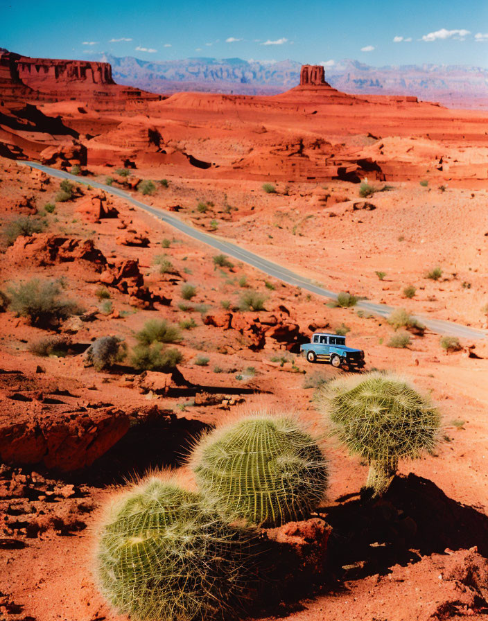 Vehicle traveling through desert landscape with red sand, rock formations, and cacti.