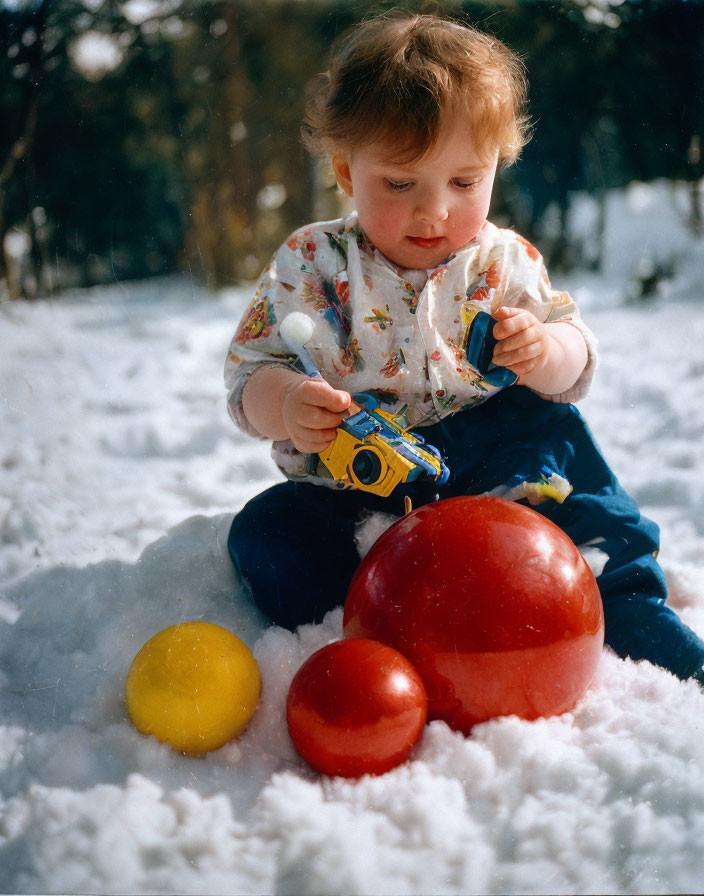 Red-haired toddler playing with colorful balls and toy truck in snow.