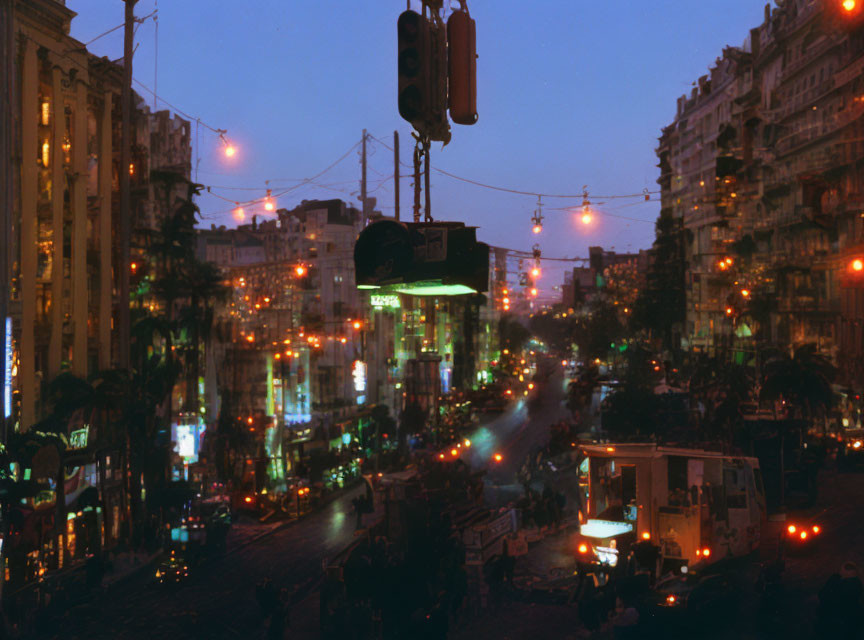 City street at dusk: cars, street lights, lit buildings, overhead cables against evening sky