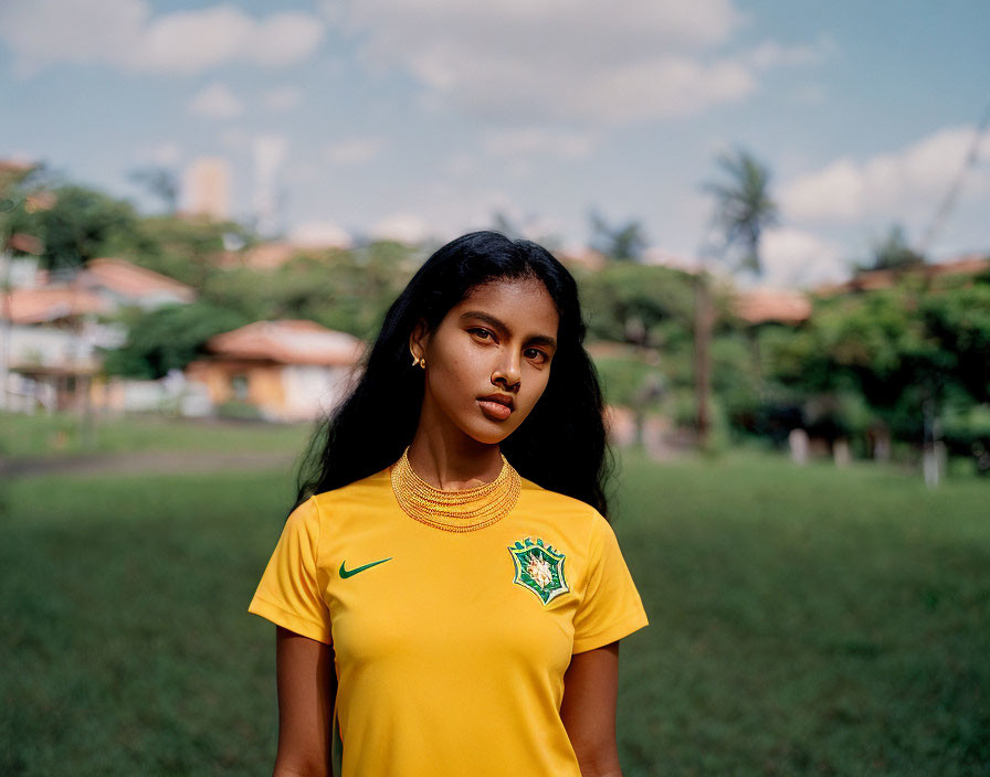 Young woman in yellow Brazil soccer jersey on grassy field with trees and buildings.