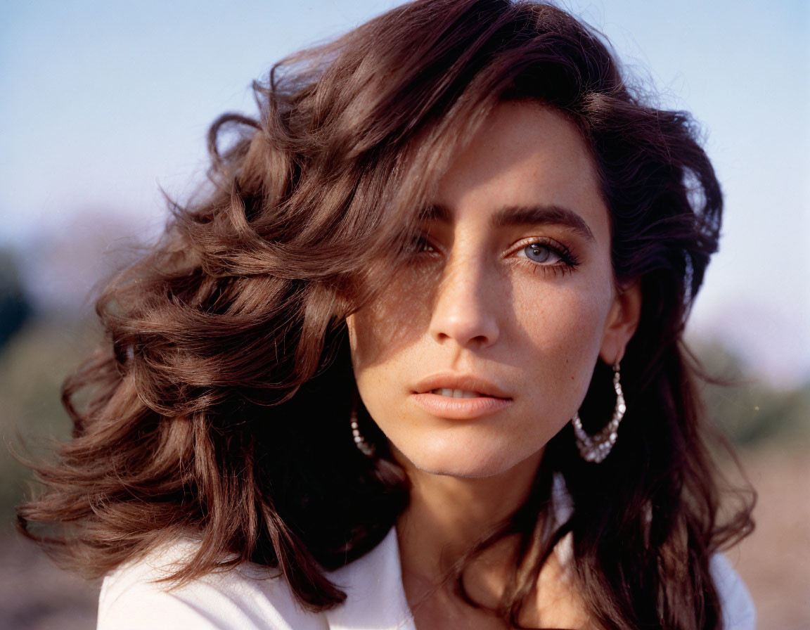 Woman with wavy brown hair and blue eyes in white top and hoop earrings gazes softly at camera