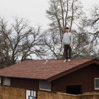 Man on wooden cabin roof surrounded by trees under overcast sky