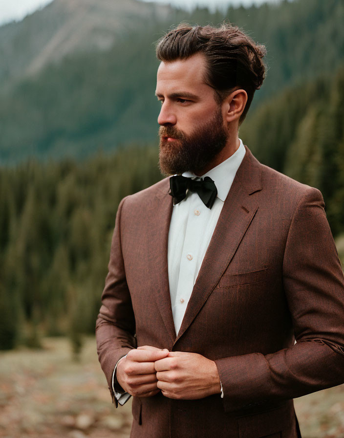 Bearded man in brown suit and bow tie outdoors with forest and mountains.