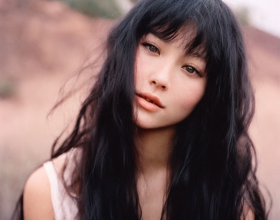 Portrait of woman with long black hair and bangs in subtle makeup, looking calmly at camera against natural