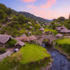 Rural cottages near stream with stone bridge in green landscape at sunset