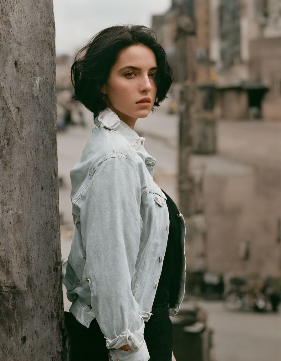 Dark-haired woman in pale denim jacket against concrete post in urban setting