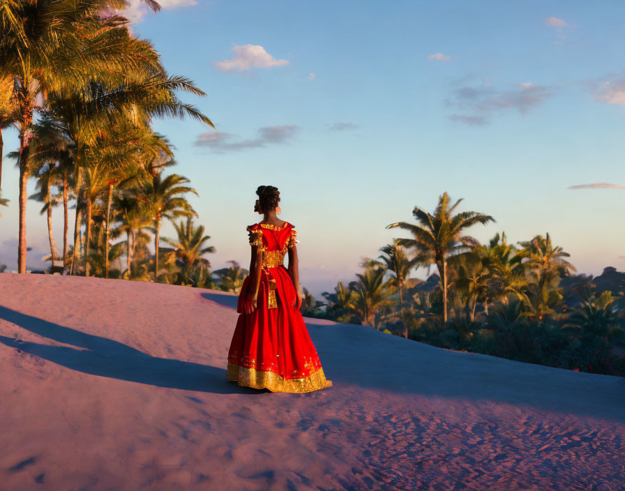 Woman in red and gold dress on sand dune at sunset with palm trees