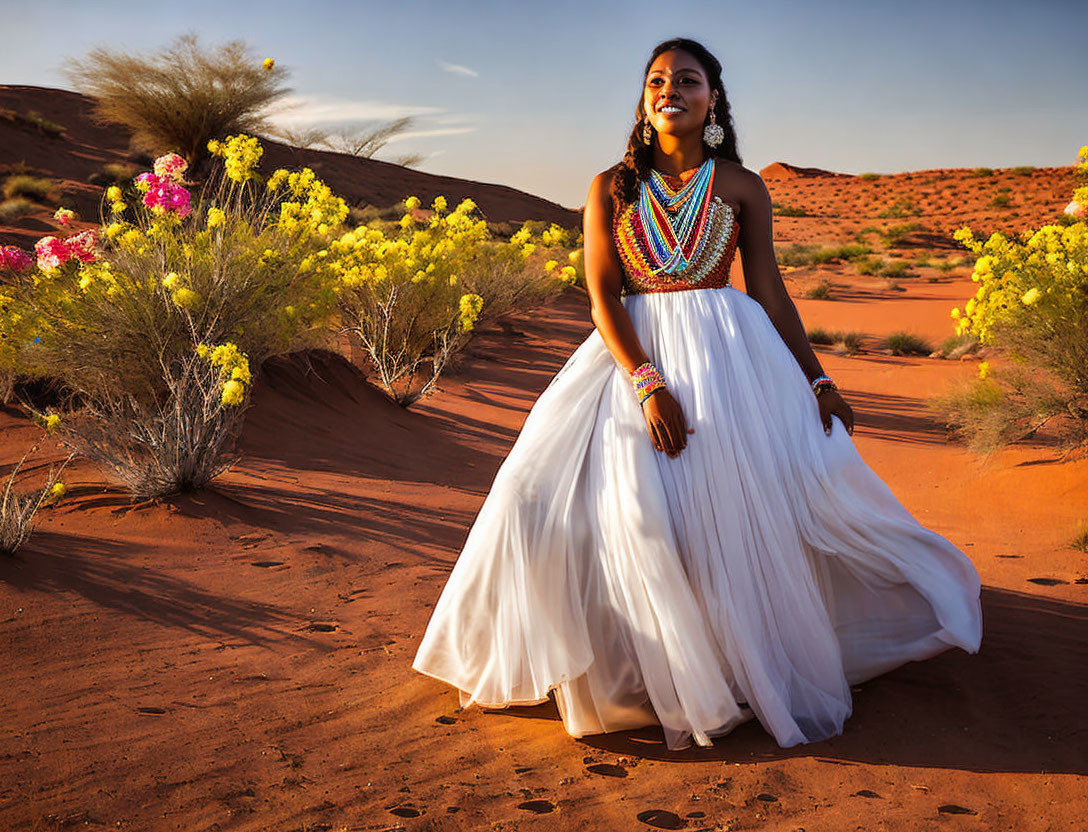 Woman in white dress with colorful jewelry poses in desert with vibrant flowers and red sand dunes.
