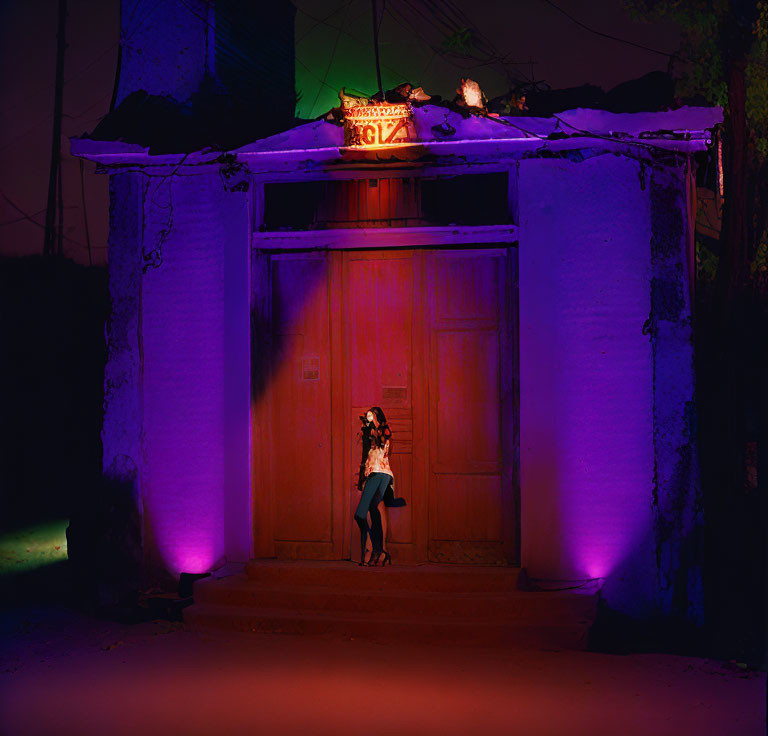 Person leaning against large wooden door under neon-lit structure at night