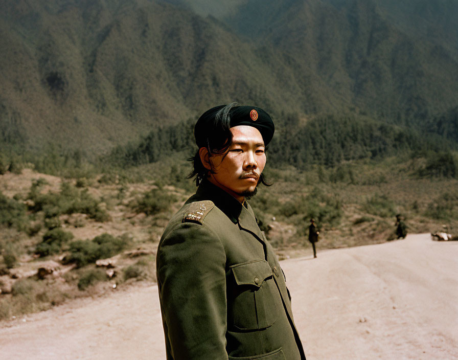 Military figure in uniform and beret on dusty road with mountains.