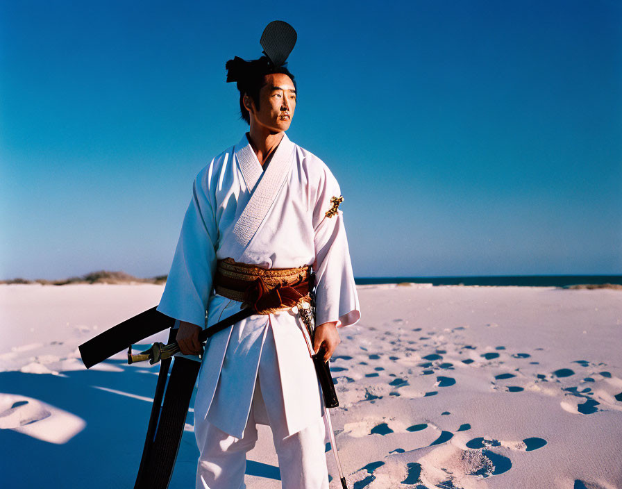Person in traditional martial arts attire with sword on sandy beach under blue sky