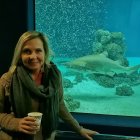 Blonde woman in green jacket smiles at table by large aquarium.