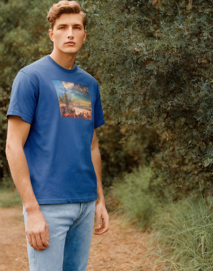 Young man in blue graphic T-shirt and light blue jeans on dirt path surrounded by green foliage