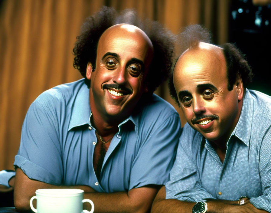 Identical bald men in blue shirts with mustaches smiling in front of warm curtain