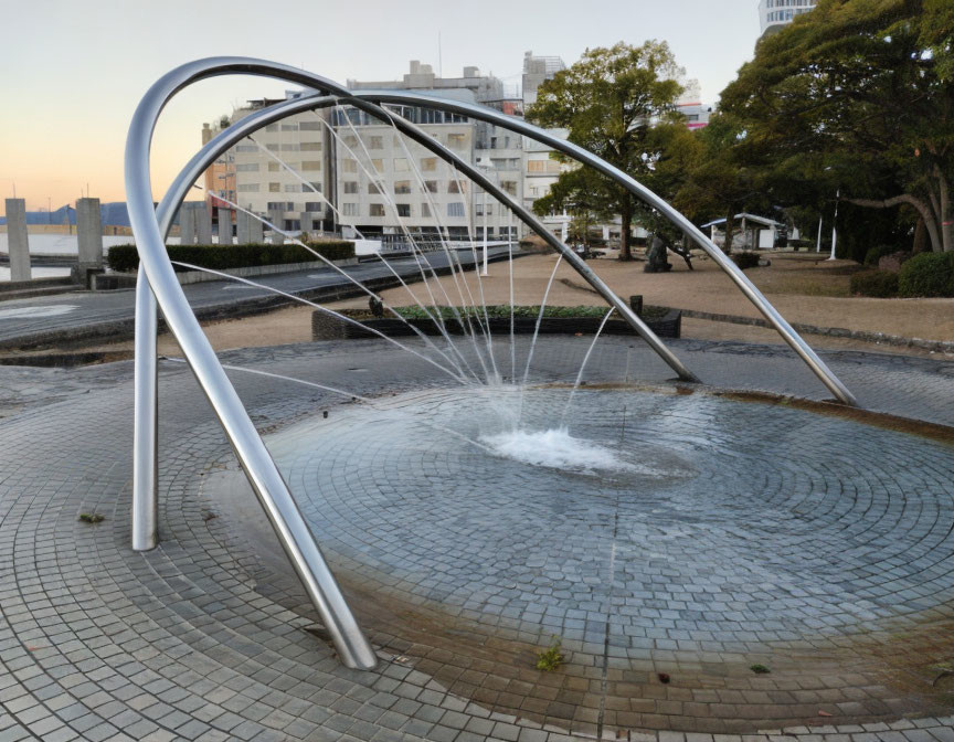 Urban fountain with arched metal structures and circular water pool amid buildings and trees.