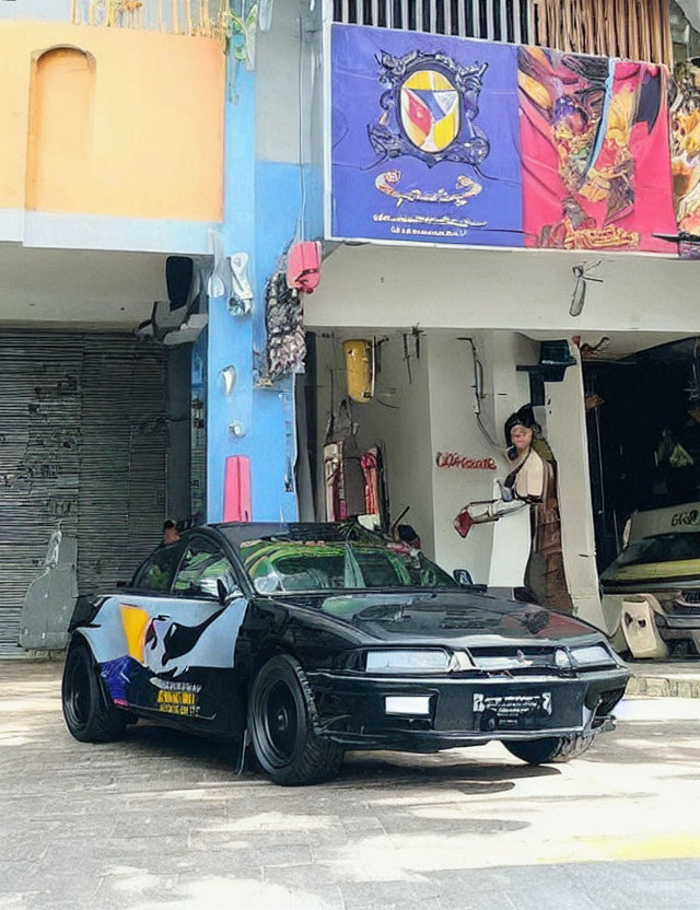 Black sports car parked in front of shop with open garage doors and person inside; colorful banners above