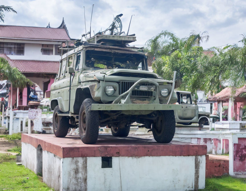 Military jeep with antenna and mounted gun on white pedestal, building in background