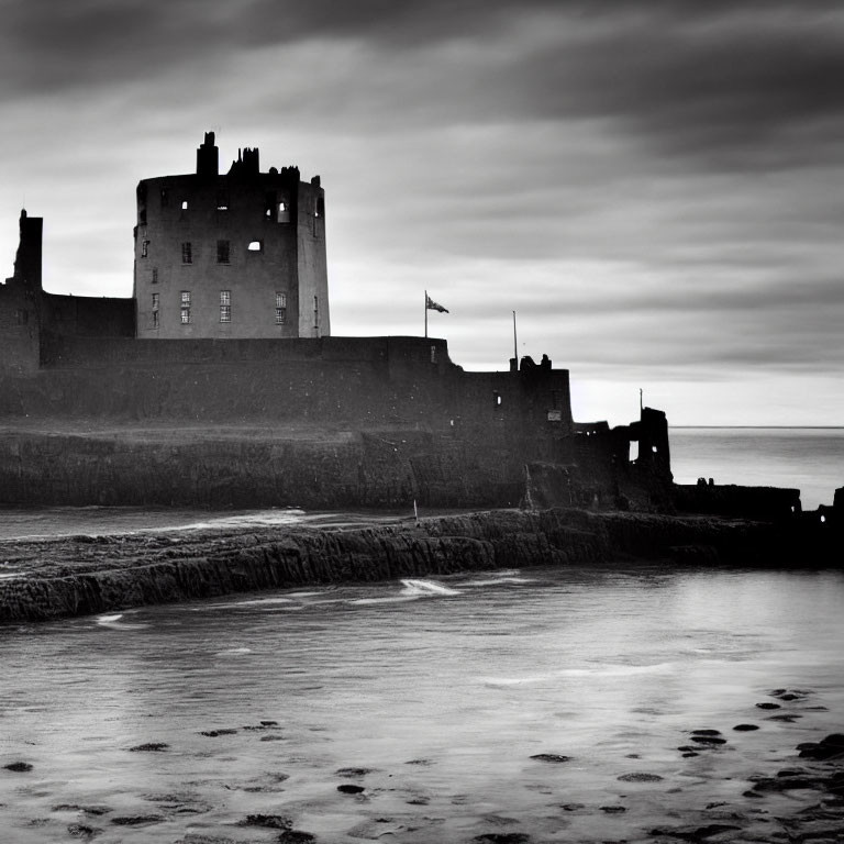 Monochrome image: ancient castle by the sea with flag, calm water, dramatic sky