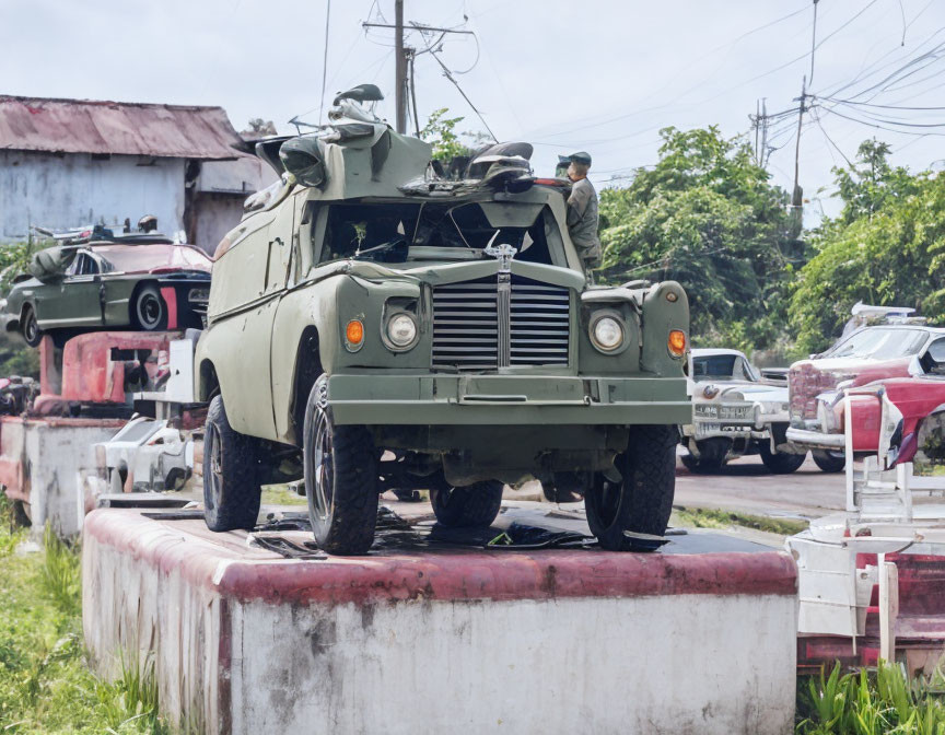 Military personnel on armored vehicle in fenced area with military and civilian vehicles.