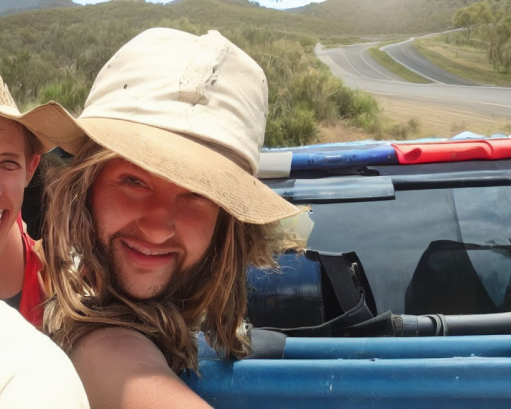 Smiling individuals in wide-brimmed hat taking selfie by blue vehicle on grassland road