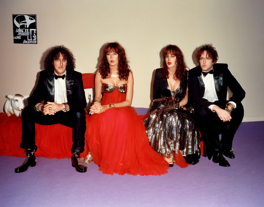 Group of four people in formal wear with a white goat, purple backdrop, and red seating