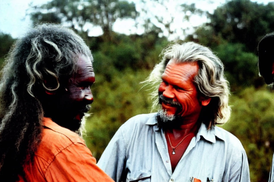 Men with painted faces in conversation: one in orange attire with traditional markings, the other in grey shirt