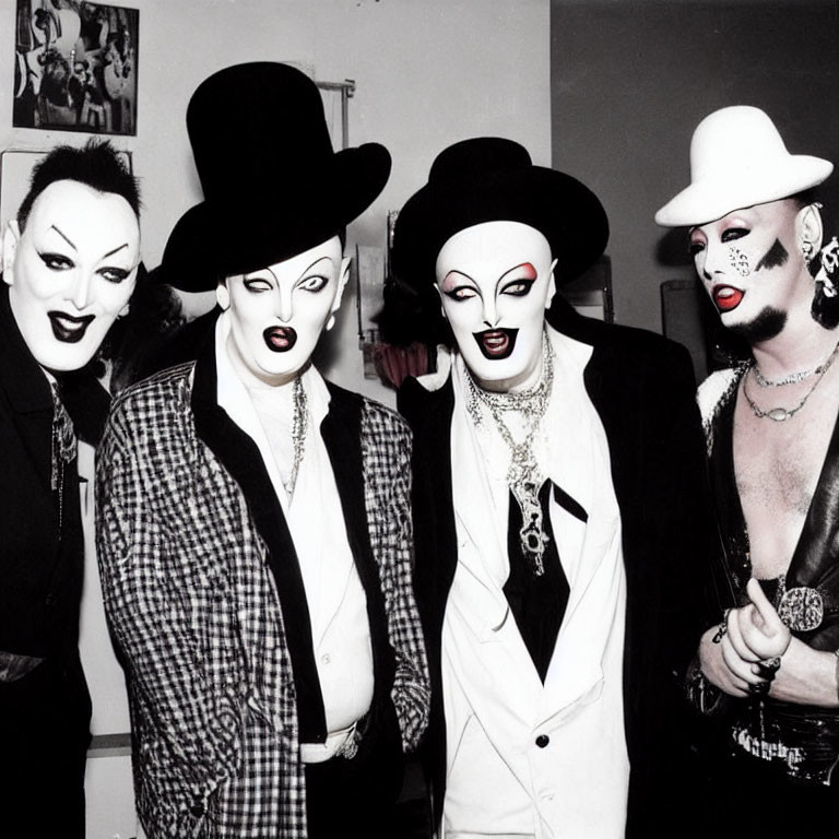 Four individuals in dramatic makeup and costumes with exaggerated facial expressions, wearing black hats and white suit elements.