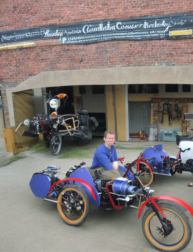 Man smiling on blue motorcycle with sidecar in front of motorcycle parts building