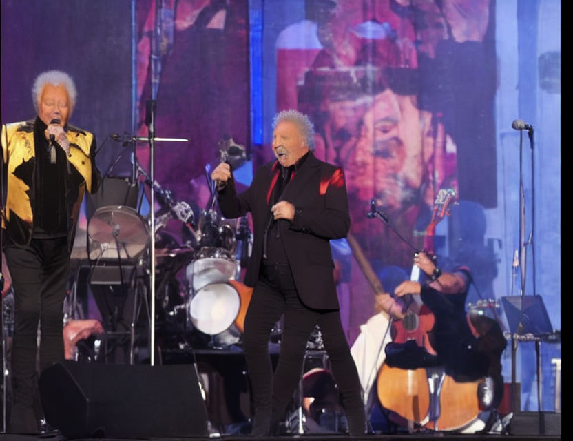 Two male singers in gold and black jackets perform on stage with a band against a backdrop showcasing their images