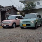 Compact pink and green cars parked by wooden house and concrete building.