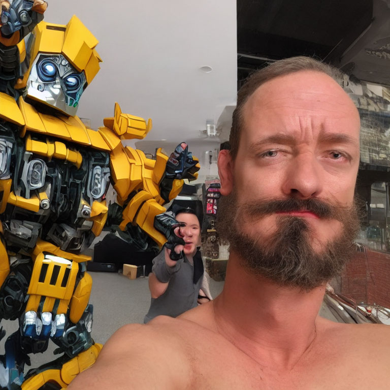 Bearded man takes selfie with Bumblebee Transformer and person aiming gun in background.