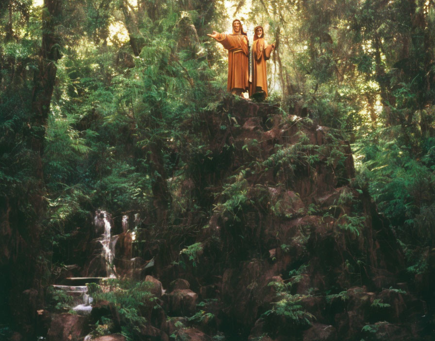 Two figures in robes on rocky outcrop in forest with waterfall.